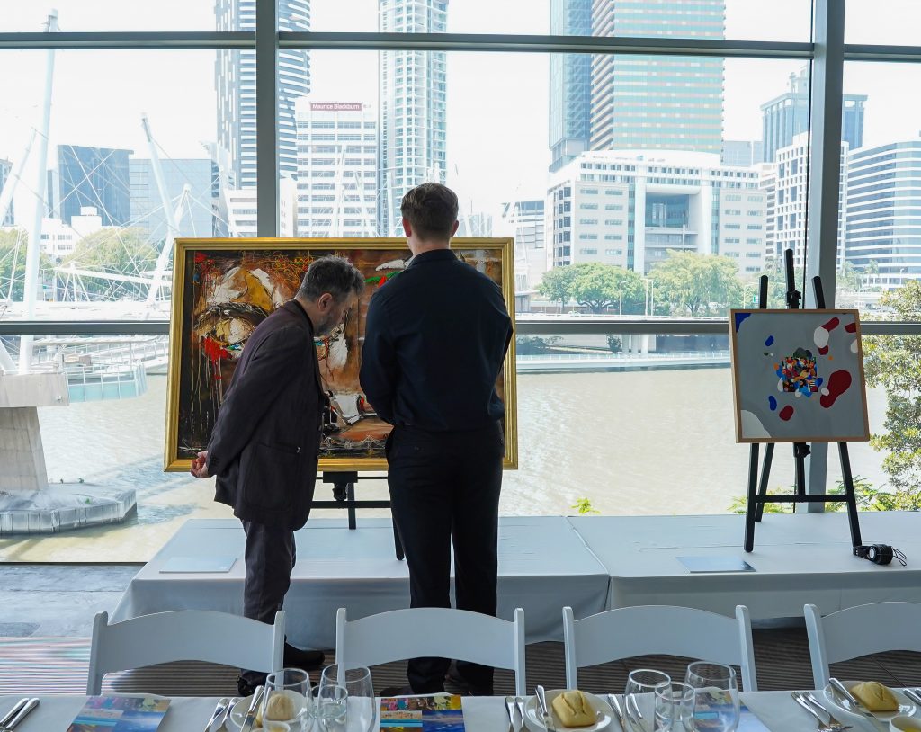 Spectators reviewing the paintings on easels against the view of Brisbane river and CBD