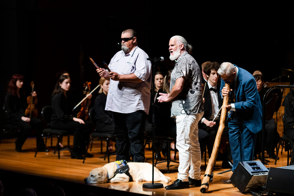 Three men stand on a stage in front of an orchestra with a guide dog at their feet, one holding clapping sticks and one playing a didgeridoo