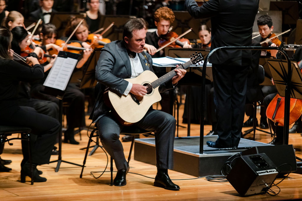 A seated man in a grey suit plays guitar in front of an orchestra's strings section
